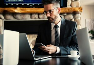 Skupiony mężczyzna w okularach i garniturze pracujący na laptopie i sprawdzający smartfona przy filiżance kawy – nowoczesne zarządzanie karierą na cvsearch.pl